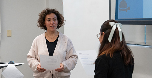 Two women stand in a classroom holding worksheets and speaking to each other.