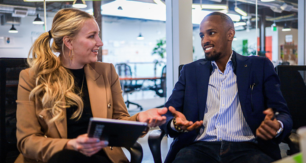 A woman and man in business attire sit together talking in a lobby area.