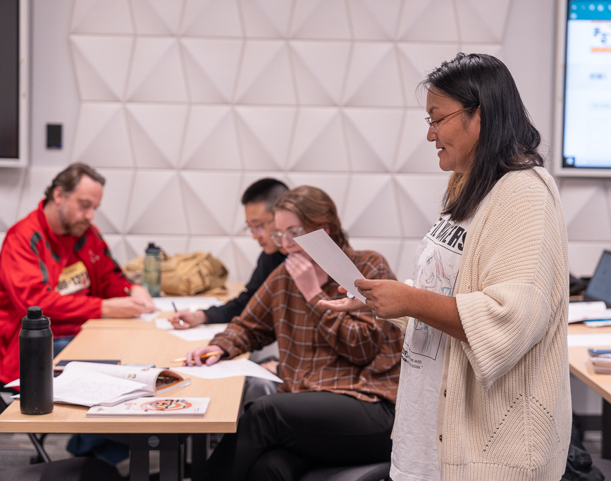 A teacher stands in front of a small class and reads from a workbook.