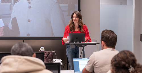 A young woman stands behind a podium teaching Spanish to adult learners.