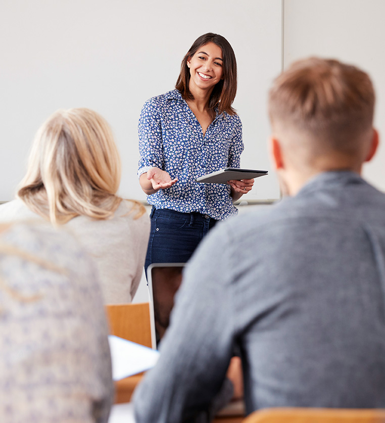 A young woman stands at the front of a classroom giving a lesson to adult learners.