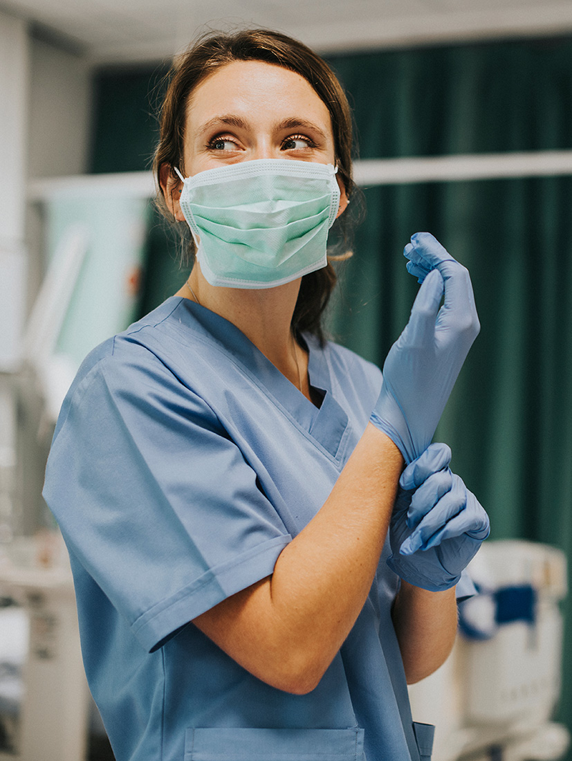 A woman in scrubs and a mask pulls on gloves in a hospital room.
