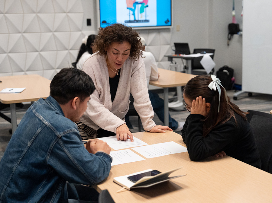 A teacher leans over a desk, talking to two students who are learning English.