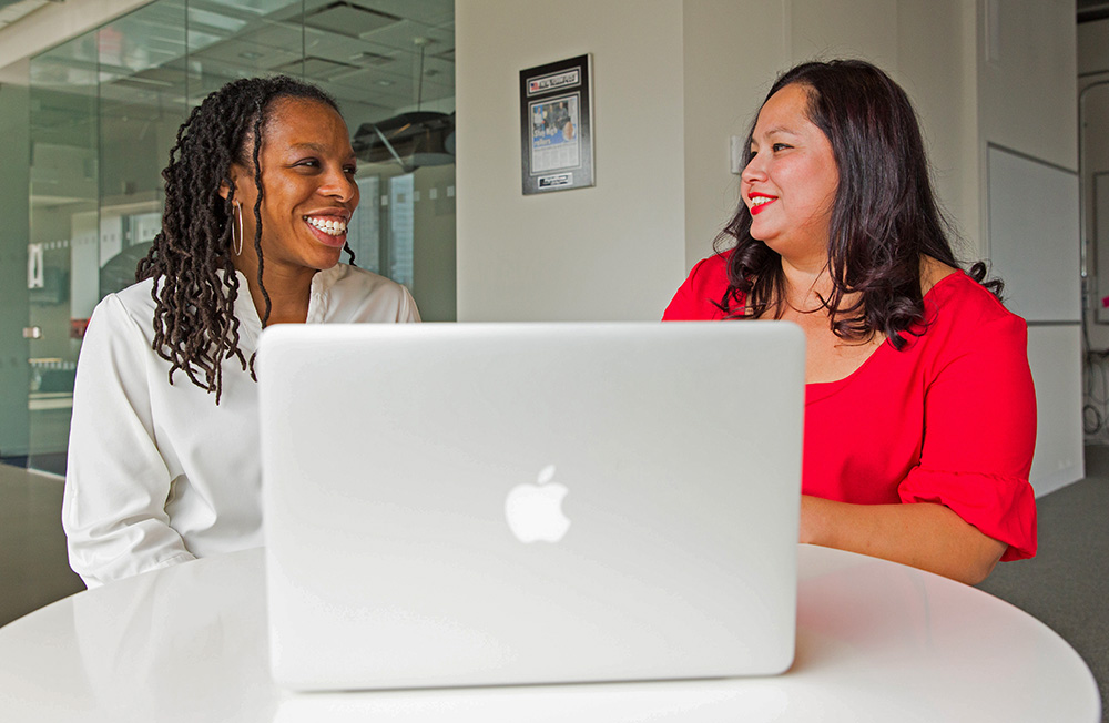 Two women sit at a small table working on a laptop.