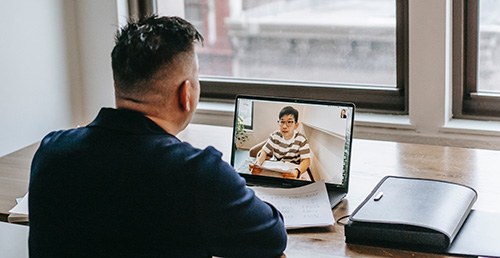 A man sits at a desk facing a window and attends a Zoom meeting.