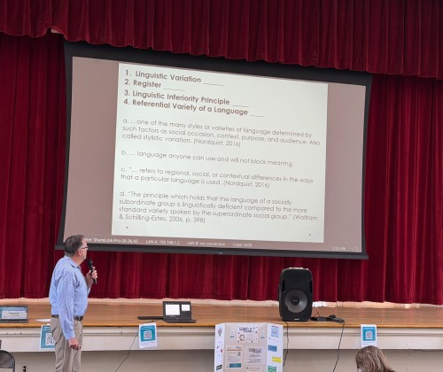 Man with microphone standing in front of a presentation screen with a slide showing