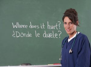 A woman in blue medical scrubs standing in front of a green blackboard with English and Spanish writing in chalk