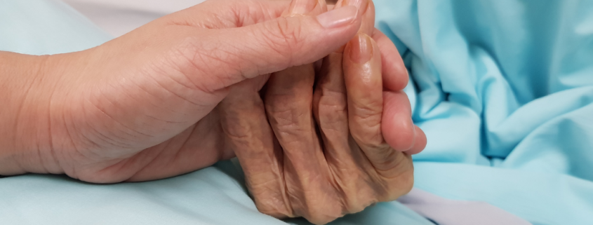 Young hand holding older, wrinkled hand on top of hospital blanket