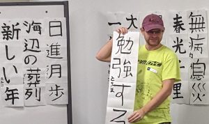 Guy standing with strip of calligraphy in front of a board covered in Japanese calligraphy