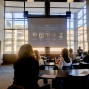 A photo from the back of the room featuring people sitting at table desks looking at a presentation screen