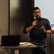 Man with dark hair and beard wearing a black shirt and holding a microphone while standing in front of a presentation screen