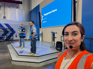 A woman with a headset and an orange vest on standing next to a stage with people doing a presentation.