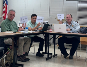 3 men sitting at tables holding up signs that say 15 in Spanish (quince)