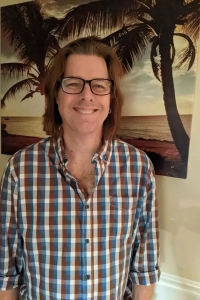 Man in glasses and button down plaid shirt standing in front of a wall painted with a palm tree.